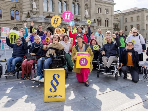 Devant un bâtiment historique à l’architecture classique, un groupe de personnes – dont plusieurs en fauteuil roulant – participe à une action publique. Elles tiennent des pancartes colorées avec des points d’exclamation et la lettre « i ». Une pancarte centrale porte le mot « INKLUSION » accompagné d’un symbole de paragraphe bleu sur fond jaune. La scène illustre une manifestation pour l’inclusion et l’accessibilité des personnes en situation de handicap, dans une atmosphère engagée et positive.