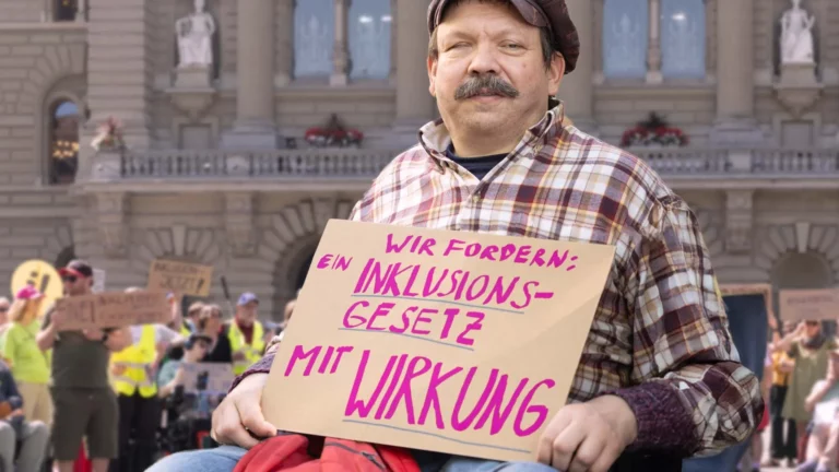 Eine Person im Rollstuhl hält ein Schild mit der Aufschrift „WIR FORDERN: EIN INKLUSIONS-GESETZ MIT WIRKUNG“. Sie befindet sich vor einem großen Regierungsgebäude, vermutlich dem Bundeshaus in Bern. Im Hintergrund sind weitere Demonstrierende mit Schildern zu sehen. Die Szene zeigt eine friedliche Protestaktion für die Rechte von Menschen mit Behinderungen und die Forderung nach einem wirksamen Inklusionsgesetz.