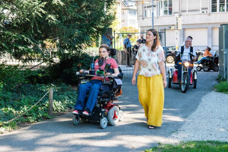 Une personne en fauteuil roulant électrique roule sur un chemin dans un parc, aidée par un accompagnateur qui marche à ses côtés. À l'arrière-plan, d'autres personnes se promènent, dont certaines utilisent également des aides à la mobilité telles que des fauteuils roulants ou des tricycles. Le chemin longe des arbres et des espaces verts avant de mener à un portail et à des bâtiments municipaux situés plus haut dans l'image. © Agile