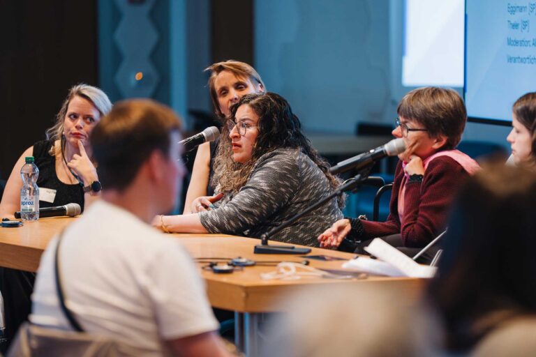 Plusieurs participant-es sont assis-es à une table sur la tribune lors de l'Assemblée des délégué-es 2026 d'Agile et discutent au micro. Au premier plan, on aperçoit le public, flou, tandis qu'à l'arrière-plan, on voit une projection avec du texte.