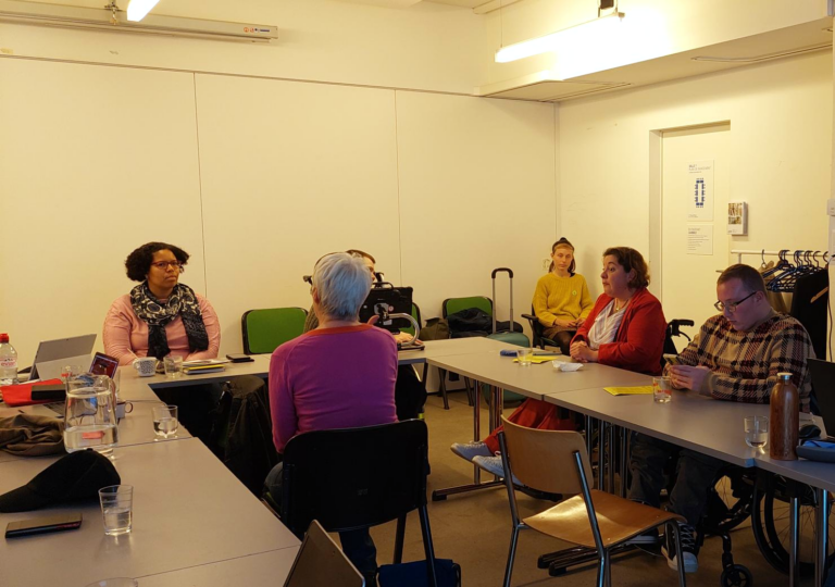 Plusieurs personnes participent à une formation autour de tables disposées en U dans une salle de réunion accessible. Des documents, des verres d’eau et des ordinateurs portables sont posés sur les tables.