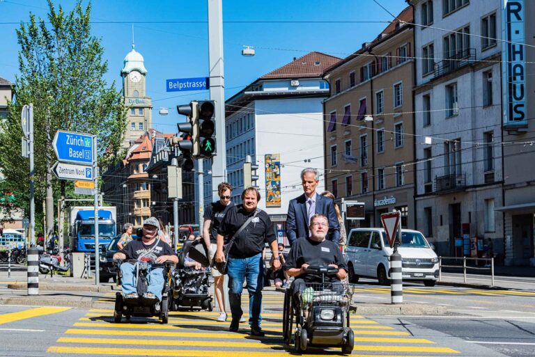 Plusieurs personnes utilisant des aides à la mobilité se déplacent de manière autonome dans l'espace public et traversent un passage piéton dans une ville suisse.
