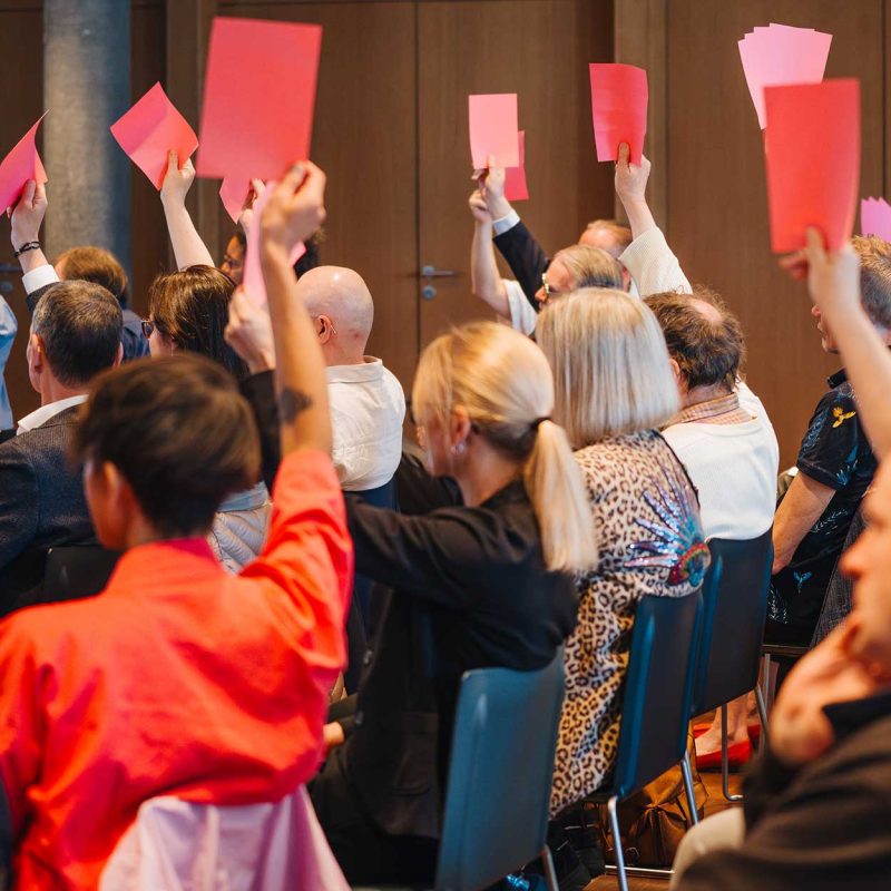 Delegierte sitzen in einem Veranstaltungsraum und stimmen ab, indem sie rote Stimmkarten hochhalten. Die Aufnahme zeigt die Abstimmungssituation während der Delegiertenversammlung 2026 von Agile aus dem Publikum heraus.
