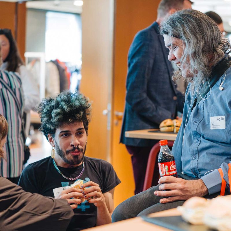 Mehrere Teilnehmende stehen und sitzen im Gesprächsbereich der Delegiertenversammlung 2026 von Agile. Im Vordergrund unterhalten sich drei Personen an einem Stehtisch mit Getränken und Snacks, im Hintergrund sind weitere Gespräche beim Networking zu sehen.