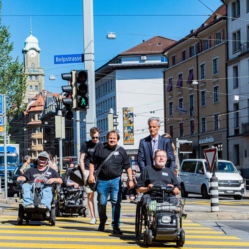 Mehrere Menschen mit Mobilitätshilfen bewegen sich selbstständig im öffentlichen Raum und überqueren einen Fussgängerstreifen in einer Schweizer Stadt.