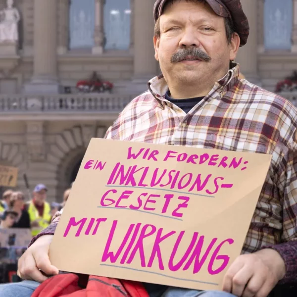 Eine Person im Rollstuhl hält ein Schild mit der Aufschrift „WIR FORDERN: EIN INKLUSIONS-GESETZ MIT WIRKUNG“. Sie befindet sich vor einem großen Regierungsgebäude, vermutlich dem Bundeshaus in Bern. Im Hintergrund sind weitere Demonstrierende mit Schildern zu sehen. Die Szene zeigt eine friedliche Protestaktion für die Rechte von Menschen mit Behinderungen und die Forderung nach einem wirksamen Inklusionsgesetz.